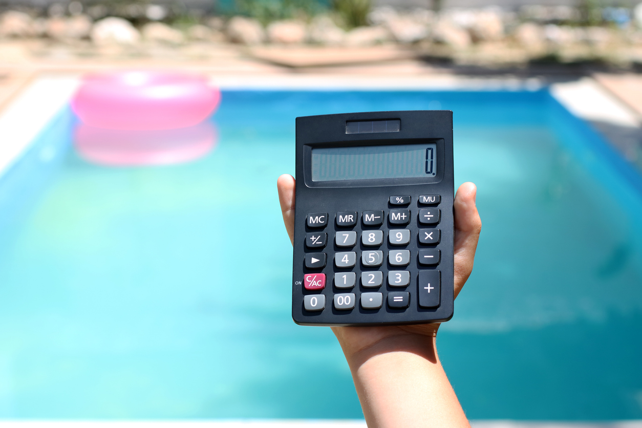 Hand holding a calculator in front of a backyard swimming pool, symbolizing pool cost estimates and budgeting in Davie, FL